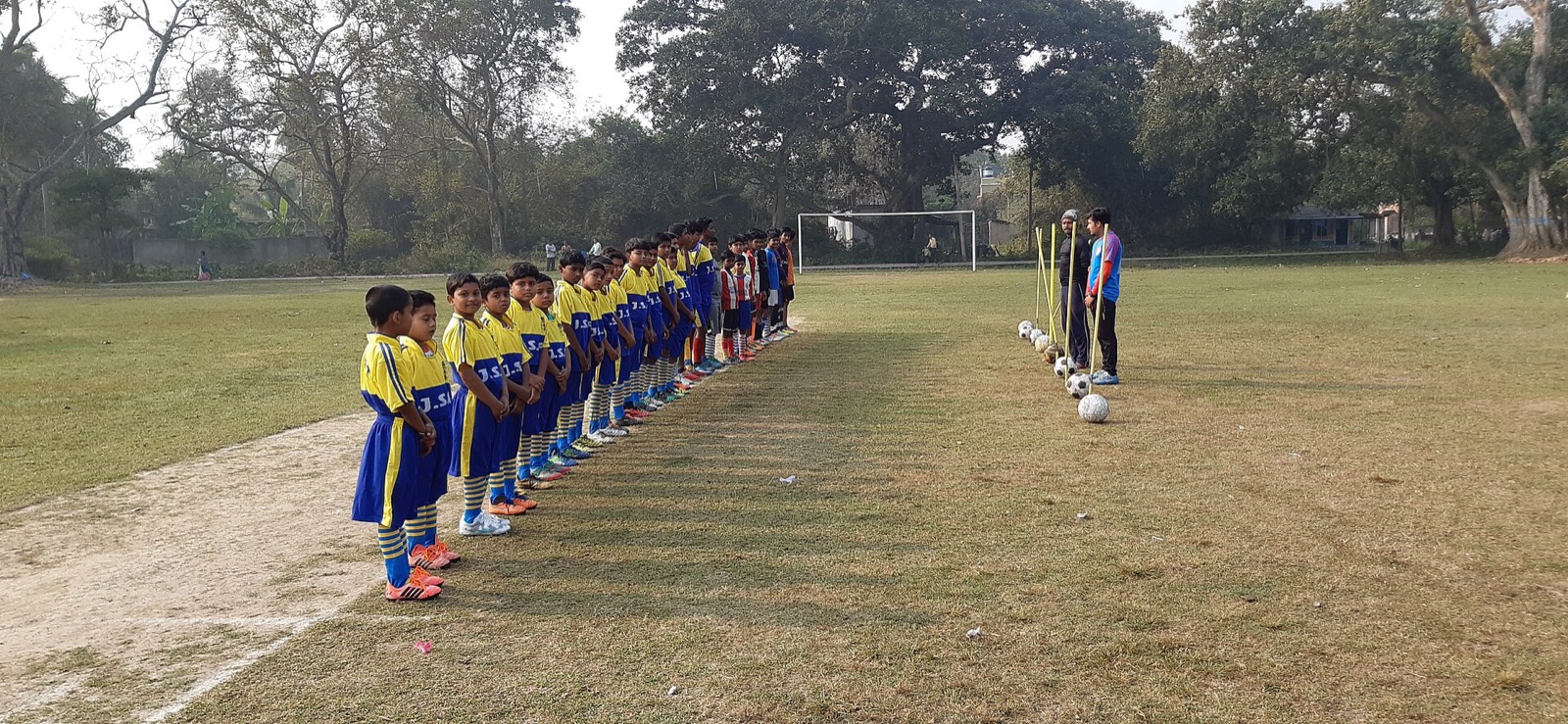 Young footballers in matching kits lined up on a ground before a training session, with coaches and balls on the right